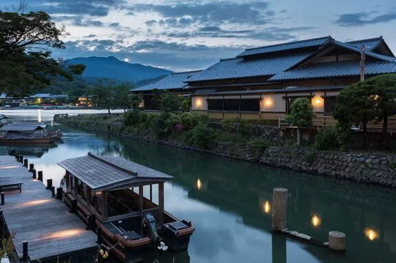 Pleasure boat and Togetsu Café old building with yellow lights at sunset, on the river bank of Saganakanoshimacho island and Arashiyama Park Nakanoshima area, Katsura River and Arashiyama hills, Ukyo Ward, Kyoto, Japan